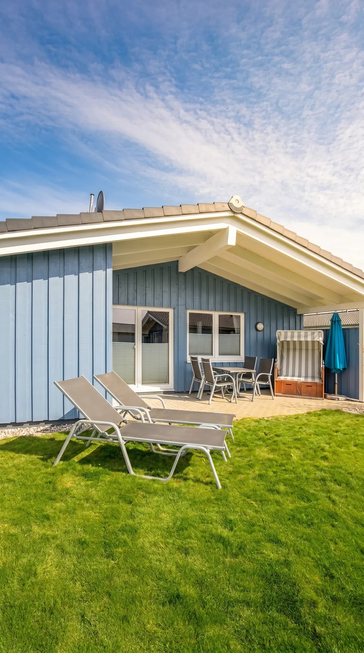 Blau verkleidetes Ferienhaus mit überdachter Terrasse, Strandkorb und zwei Sonnenliegen auf dem Rasen